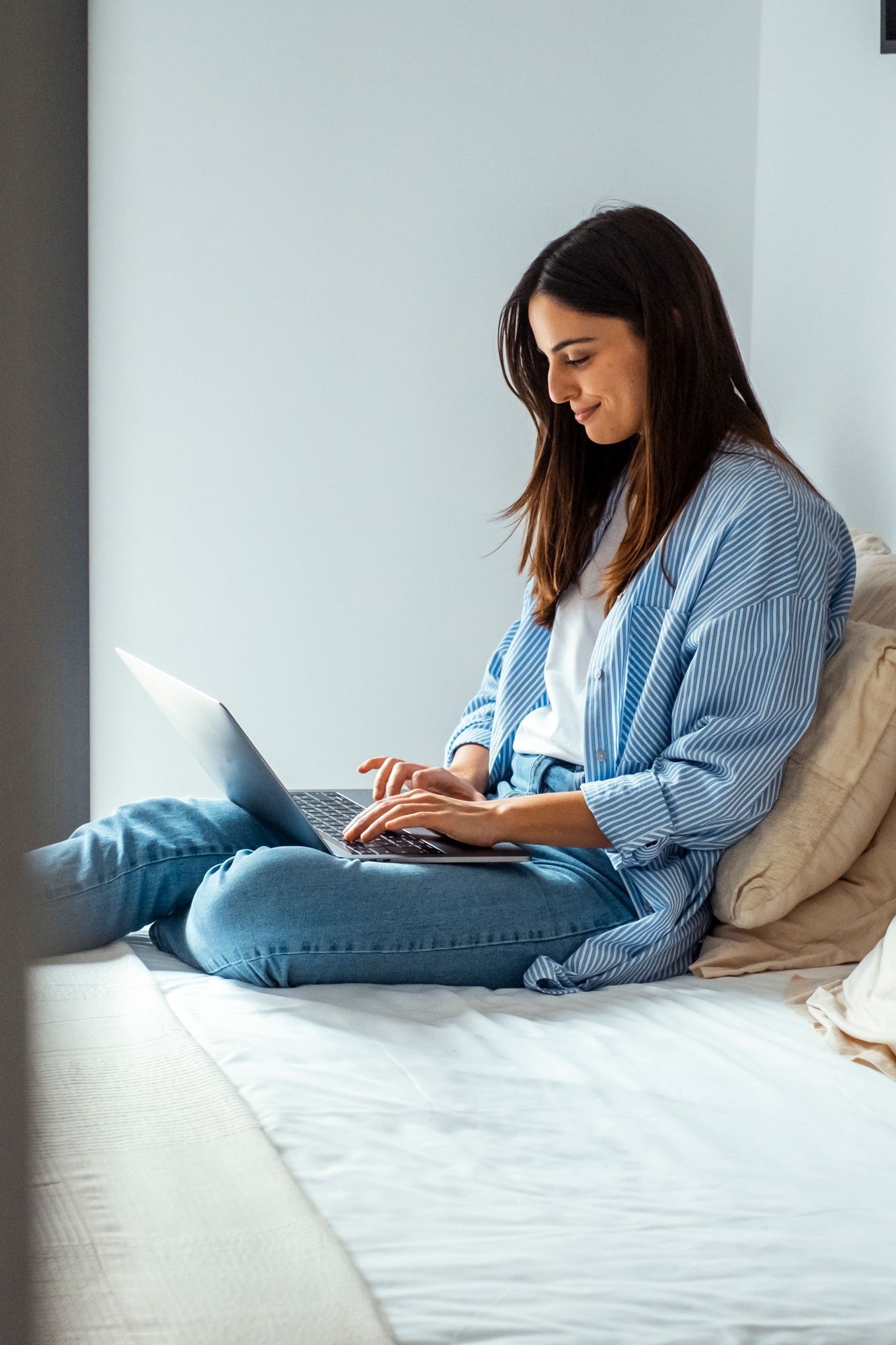 A young professional woman sits comfortably on a hotel bed, using a laptop with wireless wifi